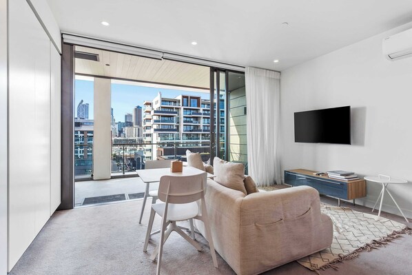 A modern living room with a sofa, TV, and dining area, featuring a city view through large windows.