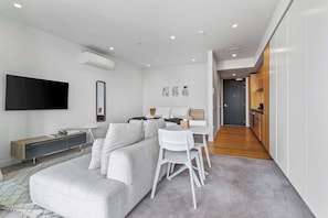 Un salon moderne comprenant un canapé, une table à manger et des chaises, un téléviseur à écran plat fixé au mur, et un coin cuisine visible à travers une porte ouverte.