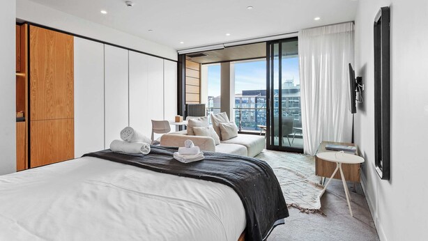 Ein modernes Hotelzimmer mit einem großen Bett, einer Sitzecke mit Sofa und Sesseln, einem kleinen Tisch und einem großen Fenster mit Blick auf die Stadt.