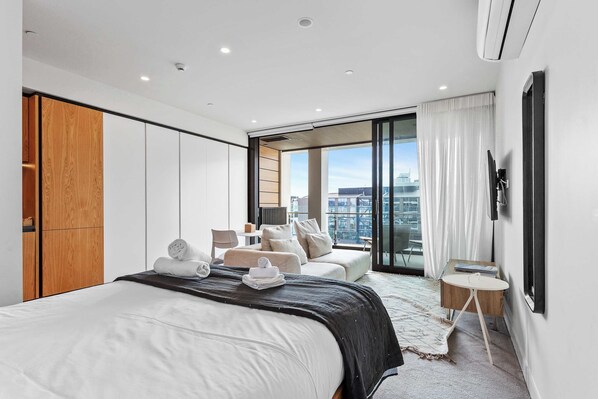 A modern hotel room with a large bed, a seating area with a sofa and chairs, a small table, and a large window offering a city view.