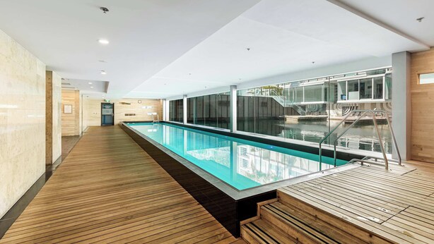 A modern indoor pool area with a wooden deck, glass walls, and a clear view of the pool. - Wynyard Viaduct Harbour 2BR+Parking: Zodiak Stays (Auckland)