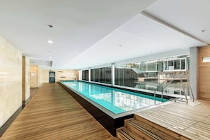 Un espace piscine intérieur moderne, doté d’une terrasse en bois, de murs en verre et offrant une vue dégagée sur la piscine.