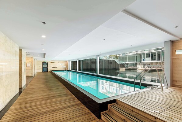 Ein moderner Poolbereich im Innenbereich mit Holzterrasse, Glaswänden und freiem Blick auf das Schwimmbecken.