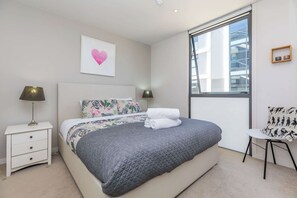 A modern bedroom with a bed, bedside lamps, a window, and a wall-mounted picture. - Wynyard Viaduct Harbour 2BR+Parking: Zodiak Stays (Auckland)