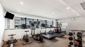 A gym with cardio machines, dumbbells, and a large window with a view of buildings. - Wynyard Viaduct Harbour 2BR+Parking: Zodiak Stays (Auckland)