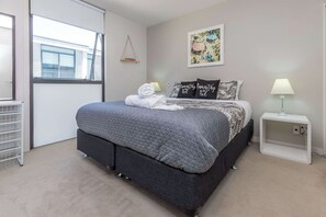 A neatly made bed with a grey quilt, a bedside lamp, and a wall-mounted shelf with books. - Wynyard Viaduct Harbour 2BR+Parking: Zodiak Stays (Auckland)