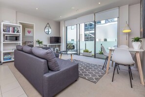 A modern living room with a grey sofa, a small round table, and a white chair. There is a bookshelf with books and a flat-screen TV.