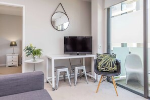 A modern living room with a flat-screen TV, a white desk, a grey sofa, a small round table with a plant, and a chair with a leaf-patterned cushion. - Wynyard Viaduct Harbour 2BR+Parking: Zodiak Stays (Auckland)