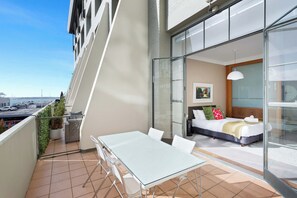 A balcony with a table and chairs, a bed with pillows, and a painting on the wall. - Lovely Bright Apt w/ Pool and Gym by Zodiak Stays (Auckland)
