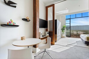 A modern hotel room with a round table, white chairs, a bed, a TV, and a large window.