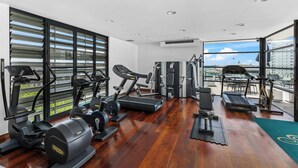 Une salle de sport équipée de divers appareils d’exercice, dotée de grandes fenêtres et offrant une vue sur la ville.