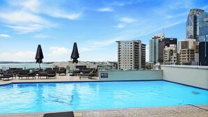 Une piscine sur le toit, dotée de chaises longues et de parasols, offrant une vue panoramique sur la ville et ses bâtiments modernes.