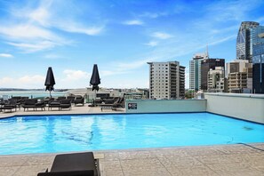 A rooftop pool with lounge chairs and umbrellas, overlooking a cityscape with modern buildings.
