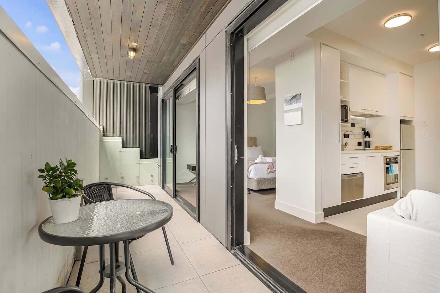 Un balcon avec une table et des chaises, une plante en pot et une porte-fenêtre coulissante donnant sur une chambre et une cuisine.