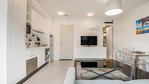Ett modernt kök med vita skåp, en inbyggd ugn och en mikrovågsugn. Det finns en matplats med ett glasbord och stolar, och en tv som är monterad på väggen.