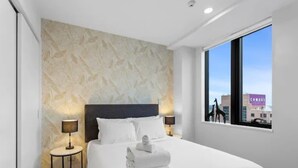 A modern hotel room with a large bed, two bedside tables, a city view, and a textured wall. - Stunning 2BR w/ Views Near Sky Tower- Zodiak Stays (Auckland)