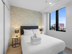 Ein modernes Hotelzimmer mit einem großen Bett, einer Wand mit Steinmusterung, zwei Nachttischlampen und Blick auf die Stadt durch ein großes Fenster.