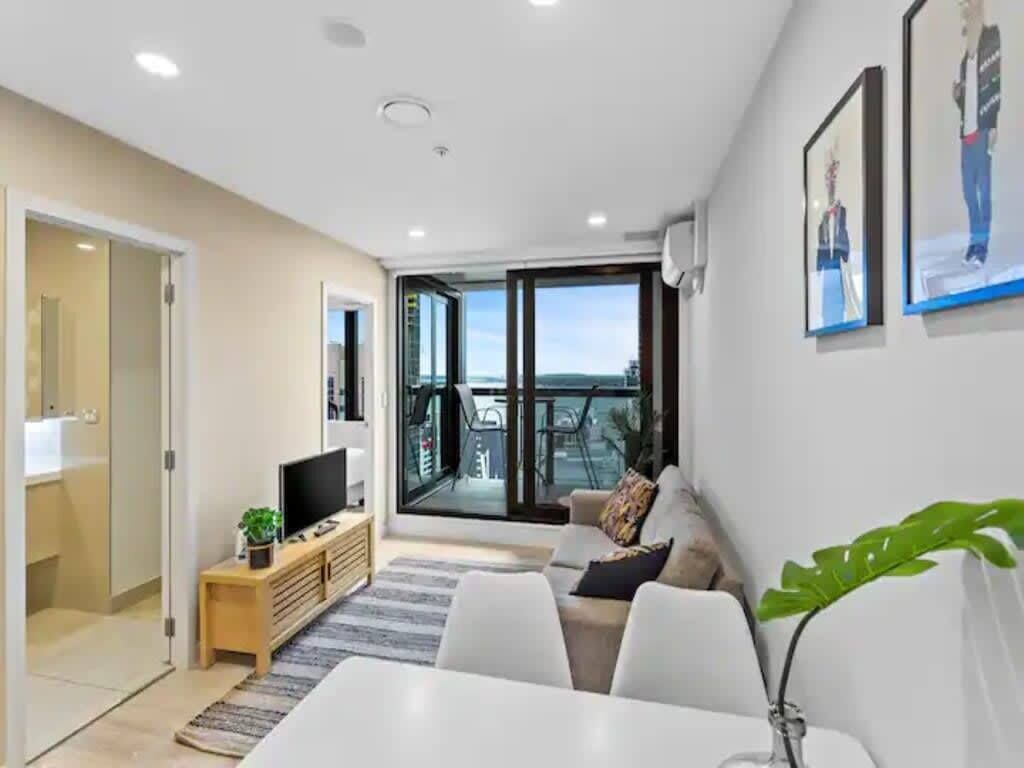 A modern living room with a sofa, a TV on a wooden stand, a plant, and a balcony with a view.