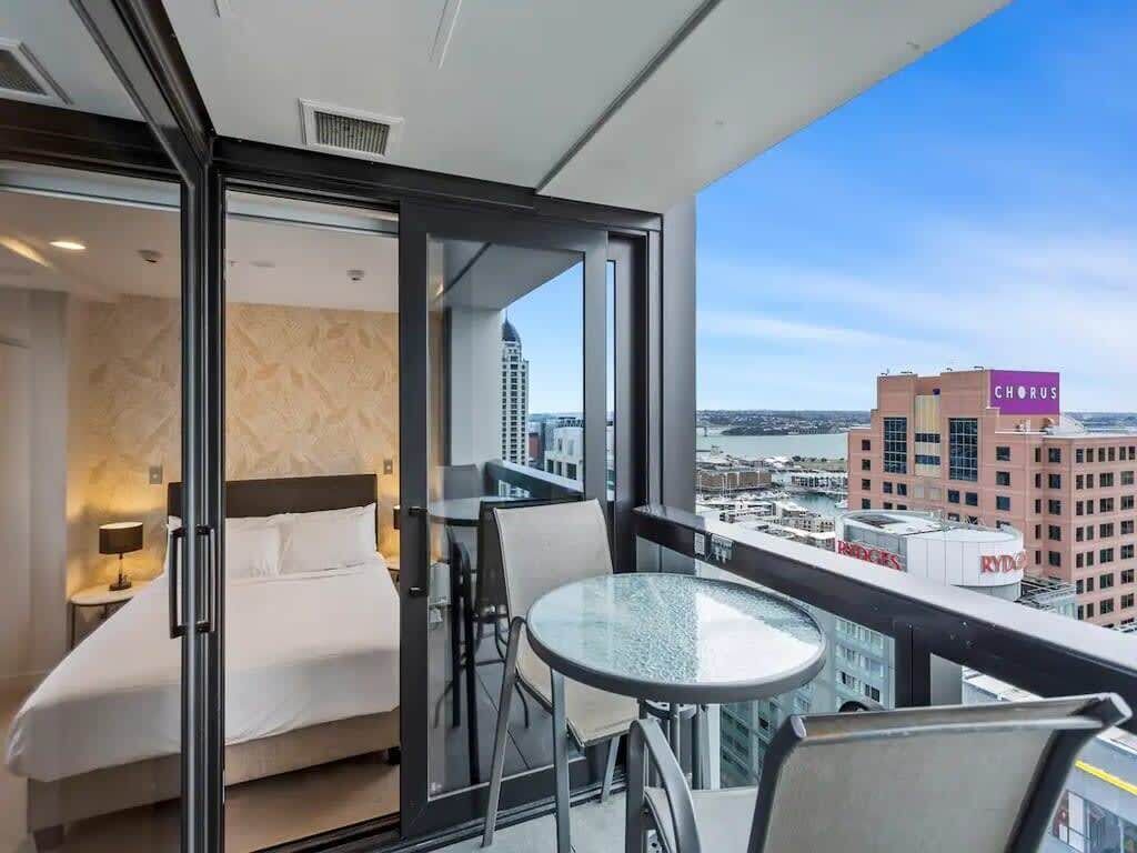 A hotel room with a bed, a glass table, and a balcony with a city view.