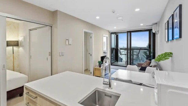 A modern kitchen with a large island, a dining area with a table and chairs, and a view through a large window. - Stunning 2BR w/ Views Near Sky Tower- Zodiak Stays (Auckland)