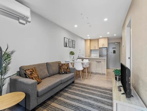 A modern living room with a sofa, a coffee table, and a television. There is a dining area with a table and chairs, and a kitchen with cabinets and a refrigerator. - Stunning 2BR w/ Views Near Sky Tower- Zodiak Stays (Auckland)