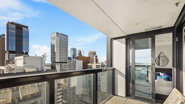 A balcony with a city view, a glass door leading to a room with a bed and a round mirror, and a view of high-rise buildings. - Sky Tower Views | 2 Bed 2 Bath CBD | Zodiak Stays (Auckland)