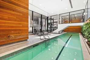 A modern indoor pool area with clear glass walls, a wooden wall on one side, and a staircase leading to an upper level. - Sky Tower Views | 2 Bed 2 Bath CBD | Zodiak Stays (Auckland)