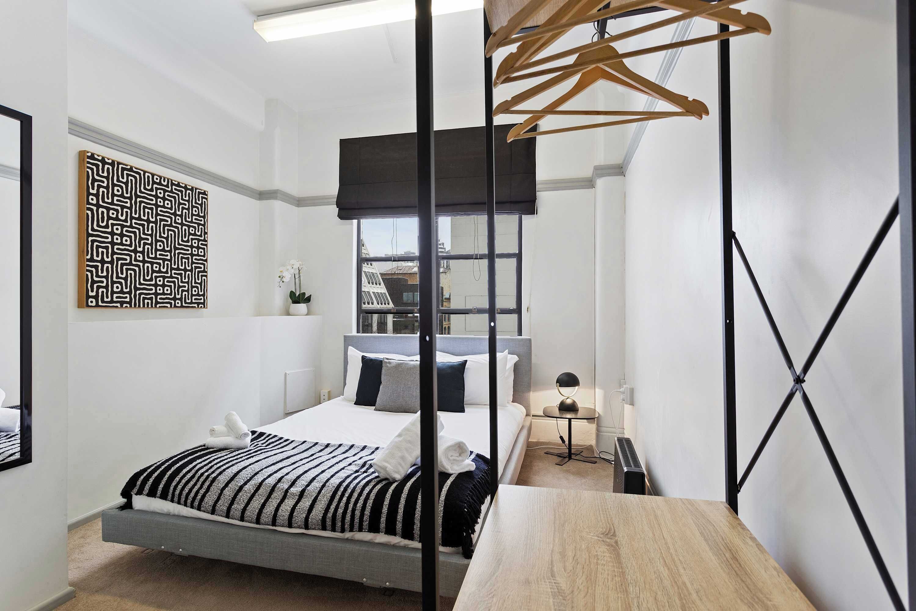 Una camera da letto moderna con un letto, un tavolo in legno, un appendiabiti e un'opera d'arte a parete.