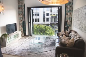 Un salon moderne avec une table basse en verre, un canapé en cuir, un téléviseur à écran plat et une grande fenêtre donnant sur la ville.
