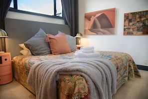 A neatly made bed with a patterned bedspread, pillows, and a folded towel. There are two bedside tables with lamps, a wall with two artworks, and a window with curtains.