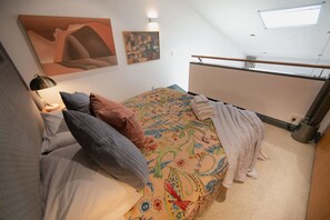 Ein Schlafzimmer mit einem Bett, Kissen, einem Nachttisch, einer Lampe, einem Fenster und einem Bild an der Wand.