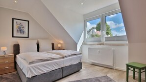 Un dormitorio con cama, lámparas de noche, un radiador, ventana con vistas y una silla verde.