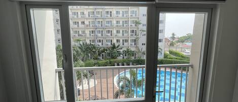Vista desde un balcón con piscina, un edificio de varios pisos y algo de vegetación al fondo.