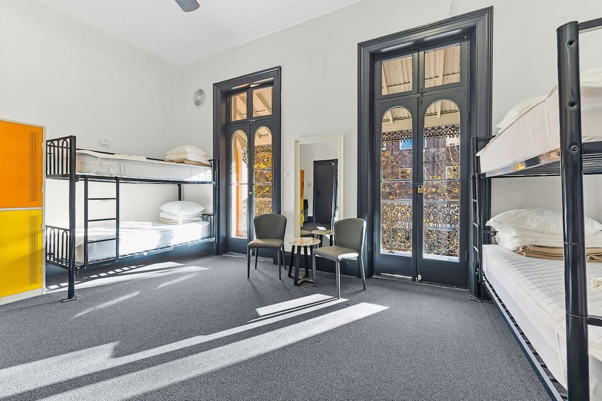 A dormitory room with bunk beds, a small table, two chairs, and a window with decorative ironwork.