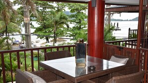 Een balkon met rieten meubels, een tafel met glazen blad en uitzicht op strand en water.
