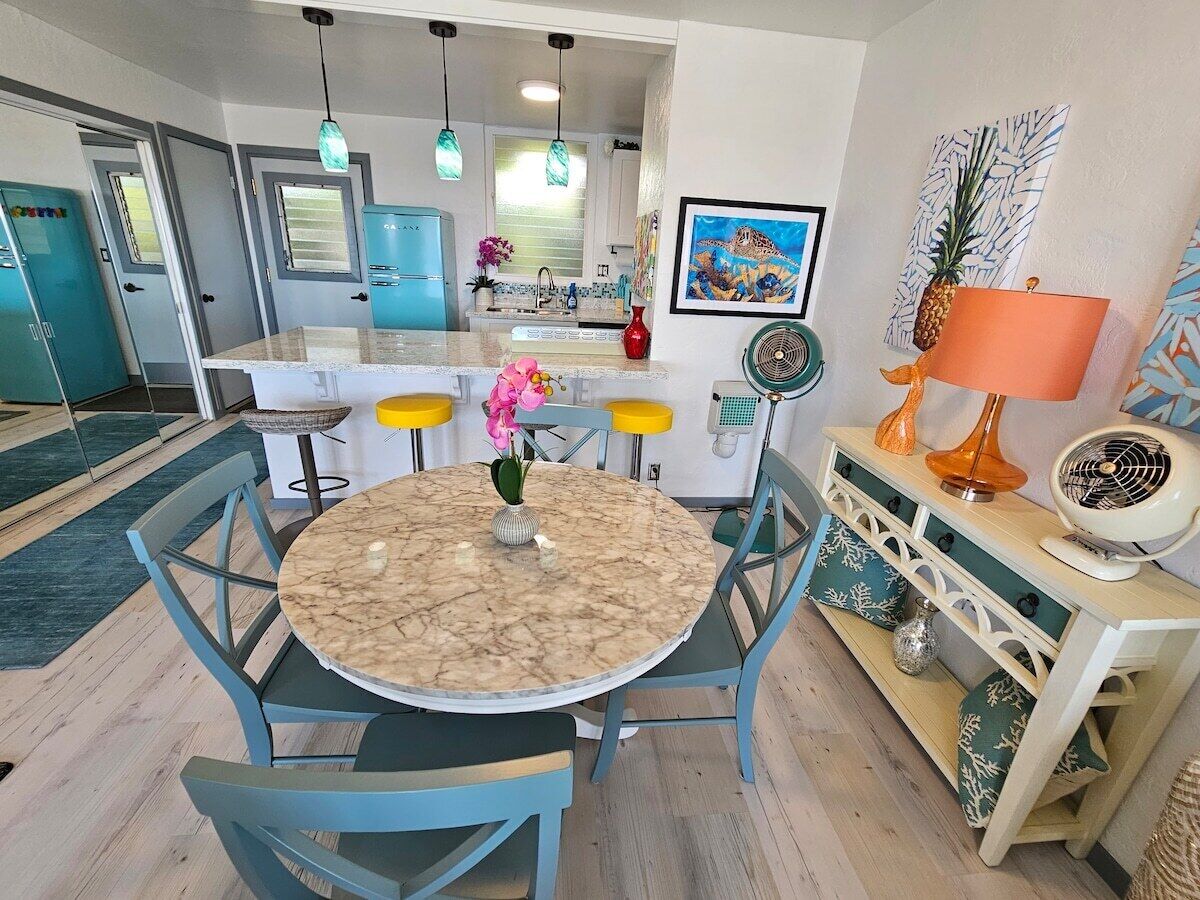 Un coin repas avec une table ronde en marbre, des chaises bleues, un petit vase avec des fleurs et un ventilateur posé sur une console en bois.