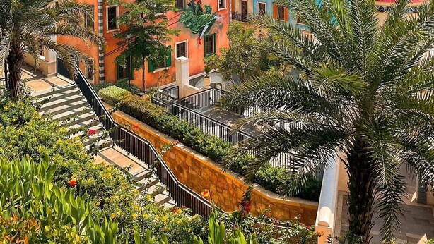 Um complexo de edifícios à beira-mar, com uma escadaria que leva a um terraço, rodeado por vegetação exuberante e um céu azul límpido.