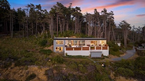 Eine moderne, zweistöckige Holzhütte mit großen Fenstern, einer Terrasse und einem Feuerkorb im Garten.