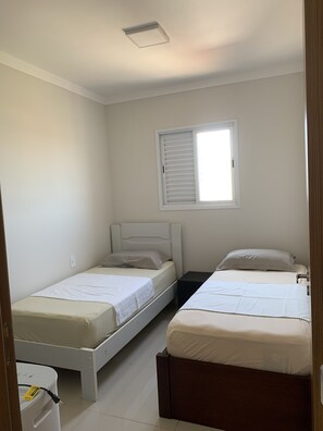 Un dormitorio con dos camas individuales, una ventana con persianas, una lámpara de techo blanca y una pared blanca.
