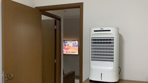 Une pièce avec un climatiseur blanc posé sur un meuble en bois, une porte donnant sur une autre pièce et un téléviseur diffusant un match de sport.