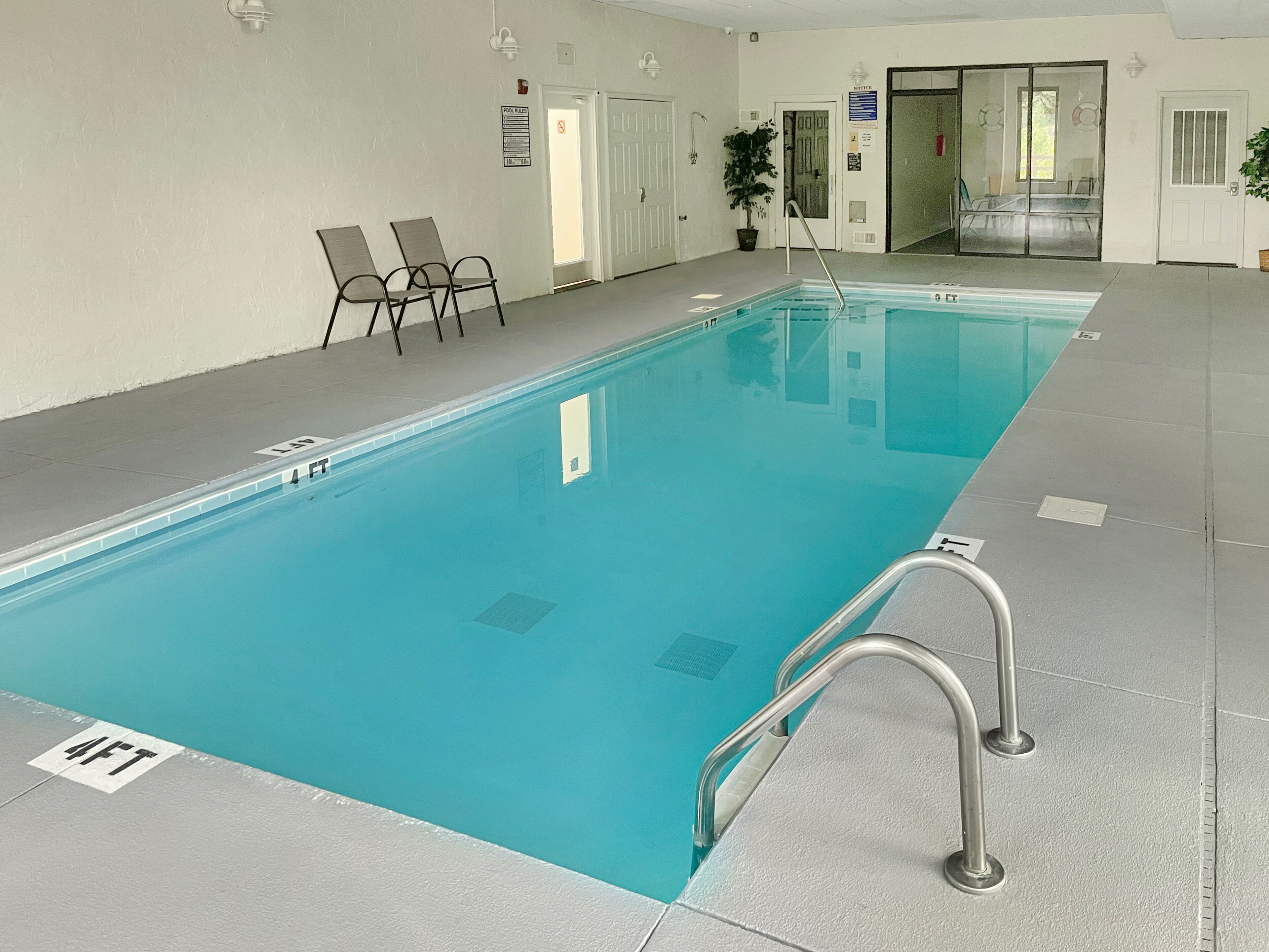 A large indoor swimming pool with a clear blue water, a metal ladder, and a few chairs near the pool.