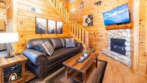 A wooden cabin interior with a sofa, a fireplace, a television, and a staircase. - Spring Special! Hot Tub, Pools, Smart TV's, Wi-Fi, Arcade, Parkway! (Pigeon Forge)