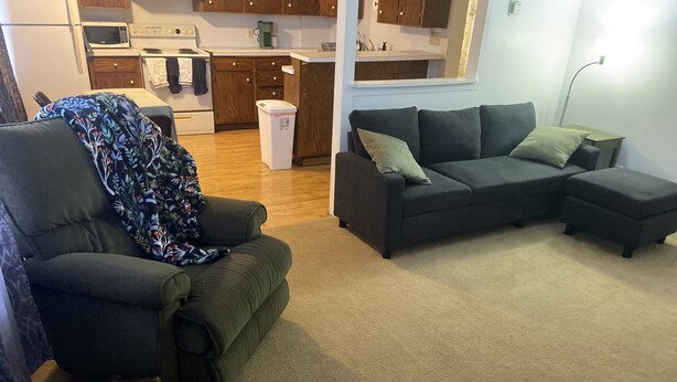 A living room with a dark green sofa, a matching armchair, and a small ottoman. A kitchen with wooden cabinets and a microwave is visible in the background. - Main Street Crosby (Crosby)