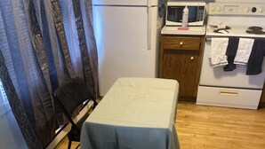 A small kitchen with a refrigerator, microwave, and stove. There is a table with a tablecloth and a chair. The room has wooden flooring and a window with curtains. - Main Street Crosby (Crosby)