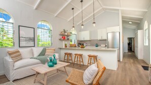 Un salon spacieux avec un canapé blanc, une table basse et un fauteuil en osier. Il y a également un coin cuisine avec un plan de travail blanc et des tabourets.