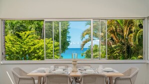 Un coin repas avec une table dressée pour quatre personnes, donnant sur une grande fenêtre qui offre une vue sur un paysage tropical et la mer.