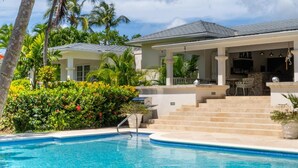 A poolside area with a covered patio, steps leading to a bar area, and lush greenery surrounding the property.