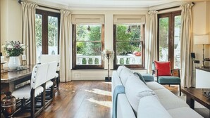 A spacious room with a large window, a sofa, a chair, and a table with a vase of flowers. - A Chelsea Retreat - 2 BR with Garden and Parking (London)
