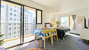 A balcony with a table and chairs, a bed, a dresser, and a large window with a view of buildings. - Sky Tower View Studio in CBD (Auckland)