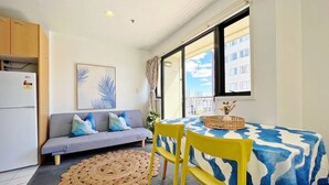 A compact living space with a sofa, a dining table with chairs, a refrigerator, and a large window providing a view of buildings. - Sky Tower View Studio in CBD (Auckland)